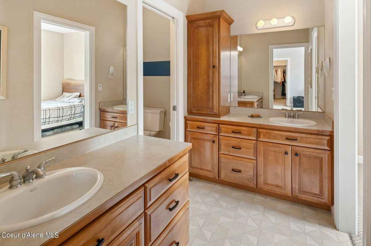 A bathroom with double sinks, wood cabinets, and a mirror is shown. A bedroom is partially visible in the background.