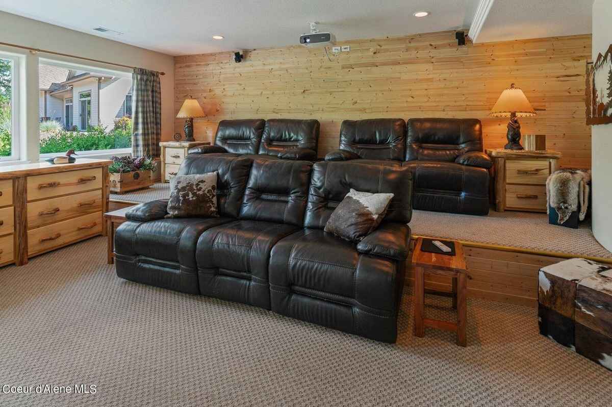 A home theater with black leather reclining seats and a wood-paneled wall is shown. There is a window with a view of trees in the background.