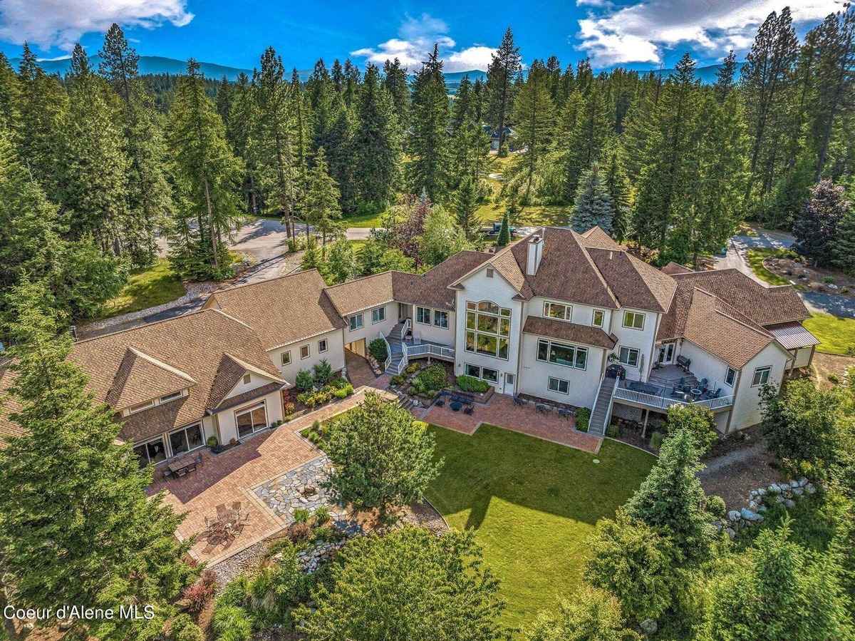 An aerial view shows a large white house with multiple additions nestled in a lush green forest, mountains visible in the background.