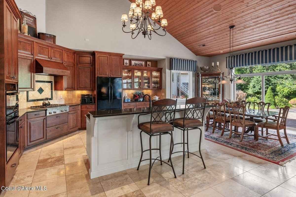 A large kitchen with dark wood cabinets, a kitchen island, and a dining area with a view of trees outside is shown.