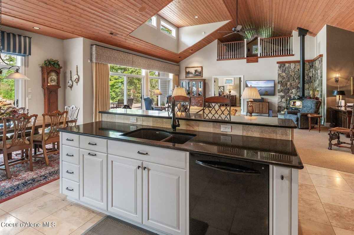 A kitchen with a large island and double sink overlooks a dining area and a spacious living room with high ceilings and a stone fireplace.