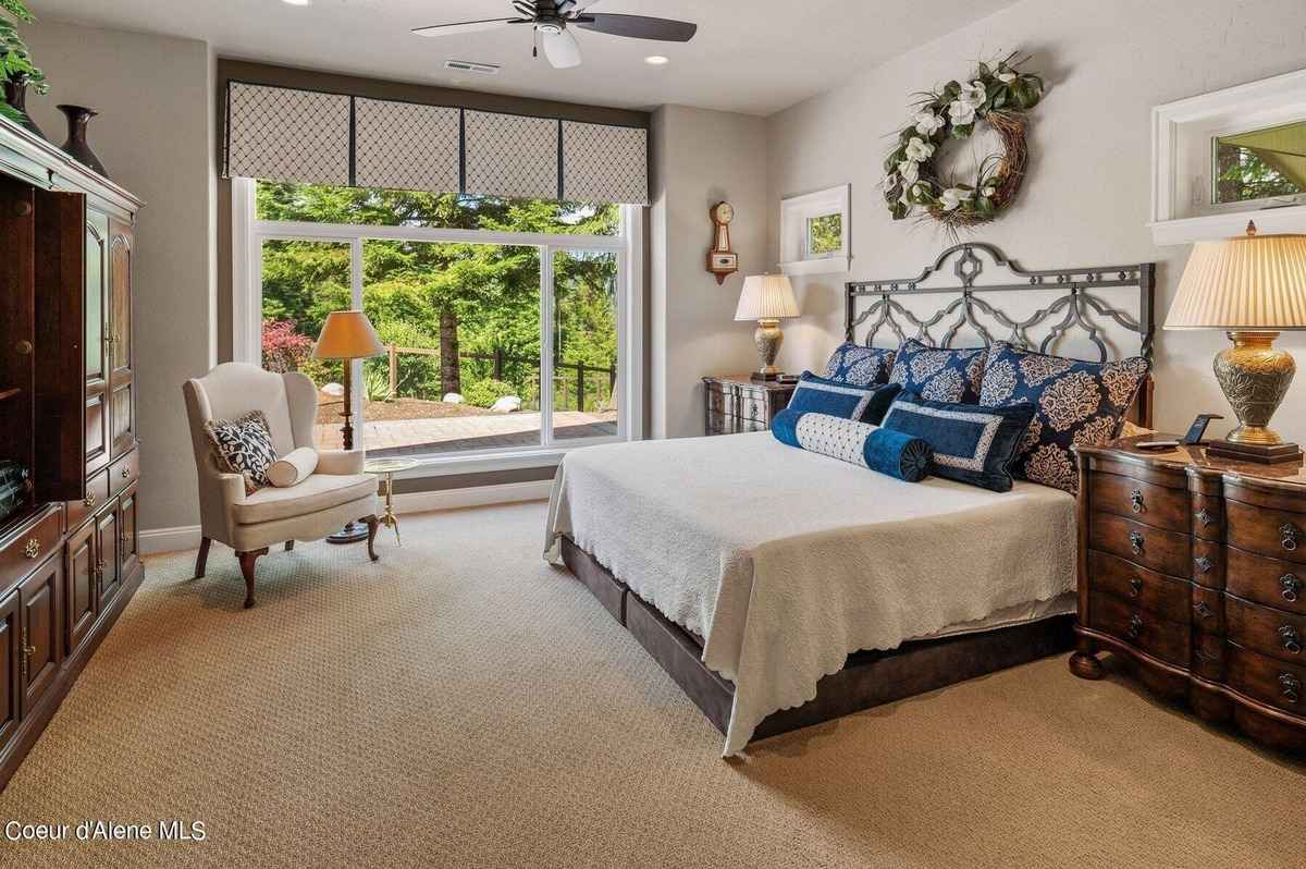 A bedroom with a large bed, a chair, and a window with a view of trees is shown. There is also a dresser and a wreath on the wall.