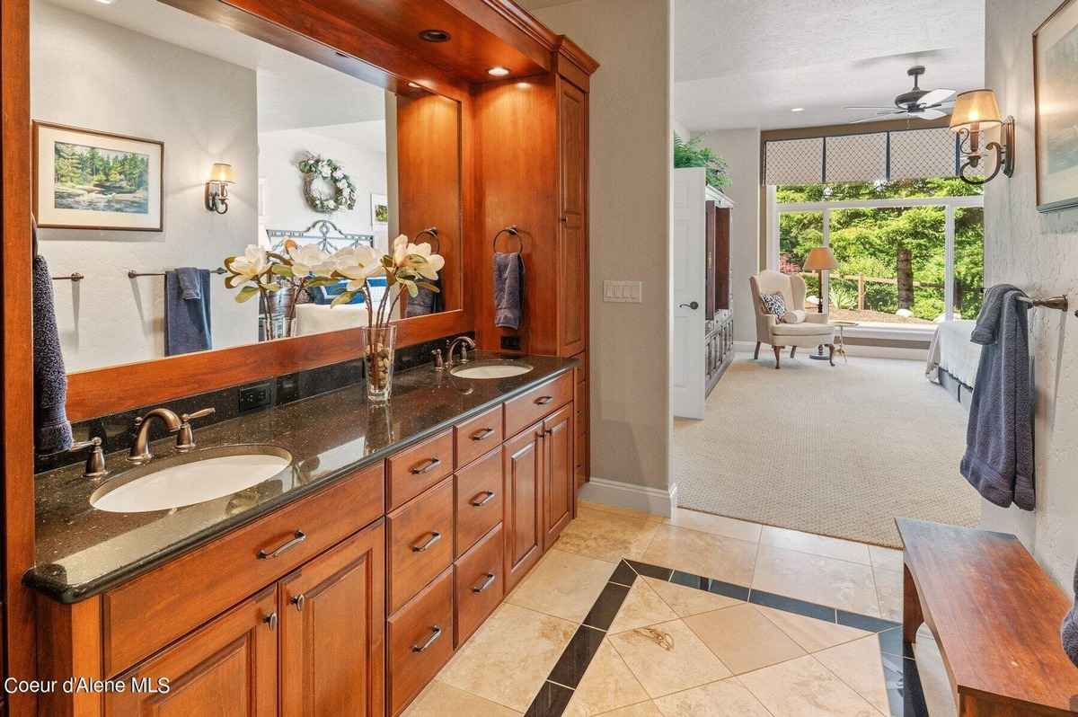 A bathroom with double sinks, dark countertops, and wood cabinets is shown. A sitting area with a window is visible in the background.