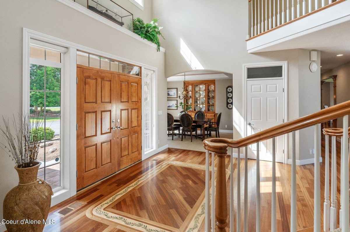 The image shows a two-story foyer with hardwood floors, a wooden staircase, and double wooden doors.