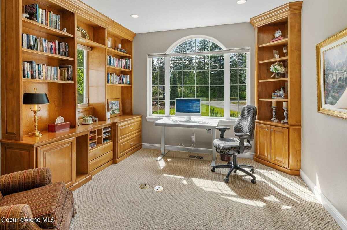 A home office with built-in bookshelves, a desk, and a chair is shown. There is a large window with a view of trees outside.