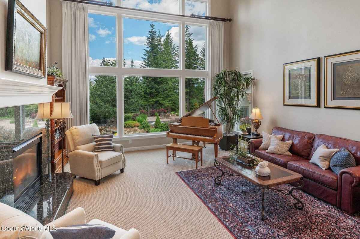 A living room with a large window, a piano, a couch, and a chair is shown. There is also a fireplace and two paintings on the wall.