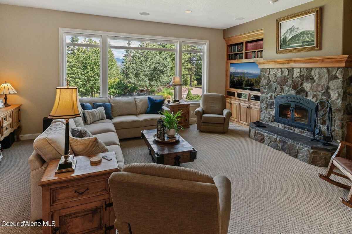 A spacious living room with a large sectional sofa, a stone fireplace, built-in shelving, and expansive windows offering a view of trees and mountains is shown.