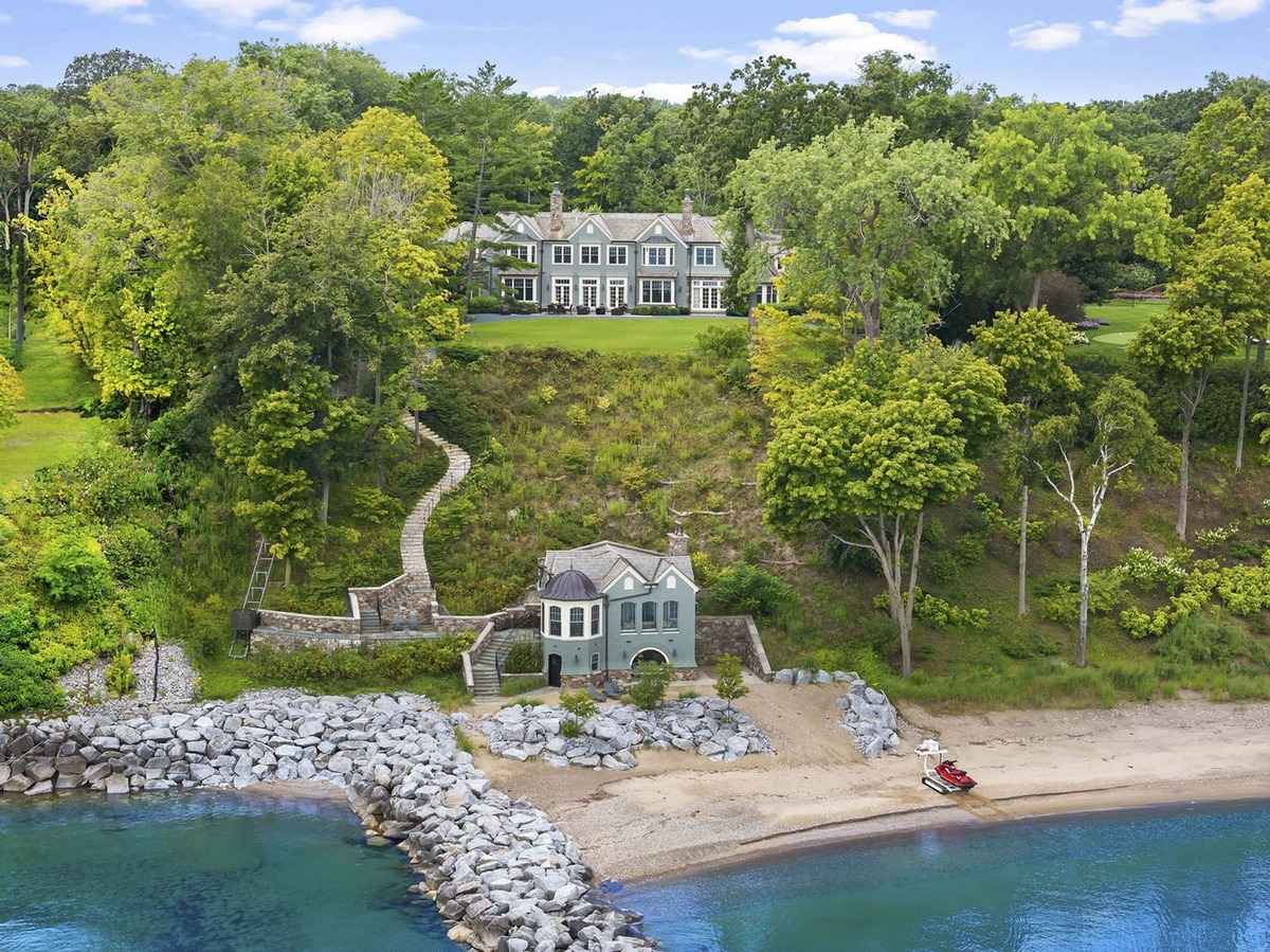 A grand lakeside estate with a main house on a hill, a stone path leading to a smaller beachfront structure, and a private beach.