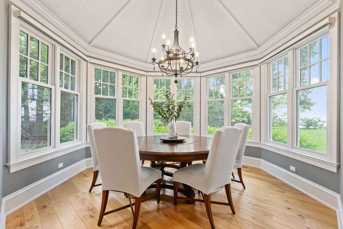 A bright dining room surrounded by large windows offering scenic views and a central chandelier.