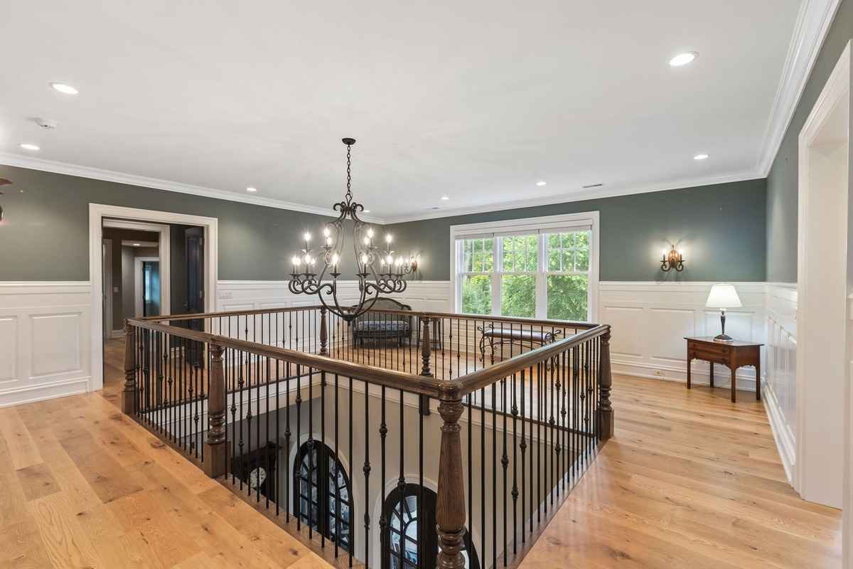 An open landing area with a large chandelier, hardwood floors, and a window view.