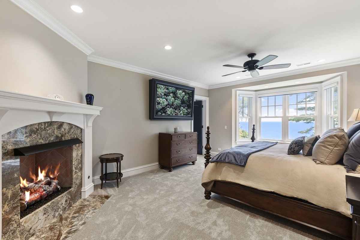 A luxurious bedroom with a fireplace, large bay windows, and serene views.