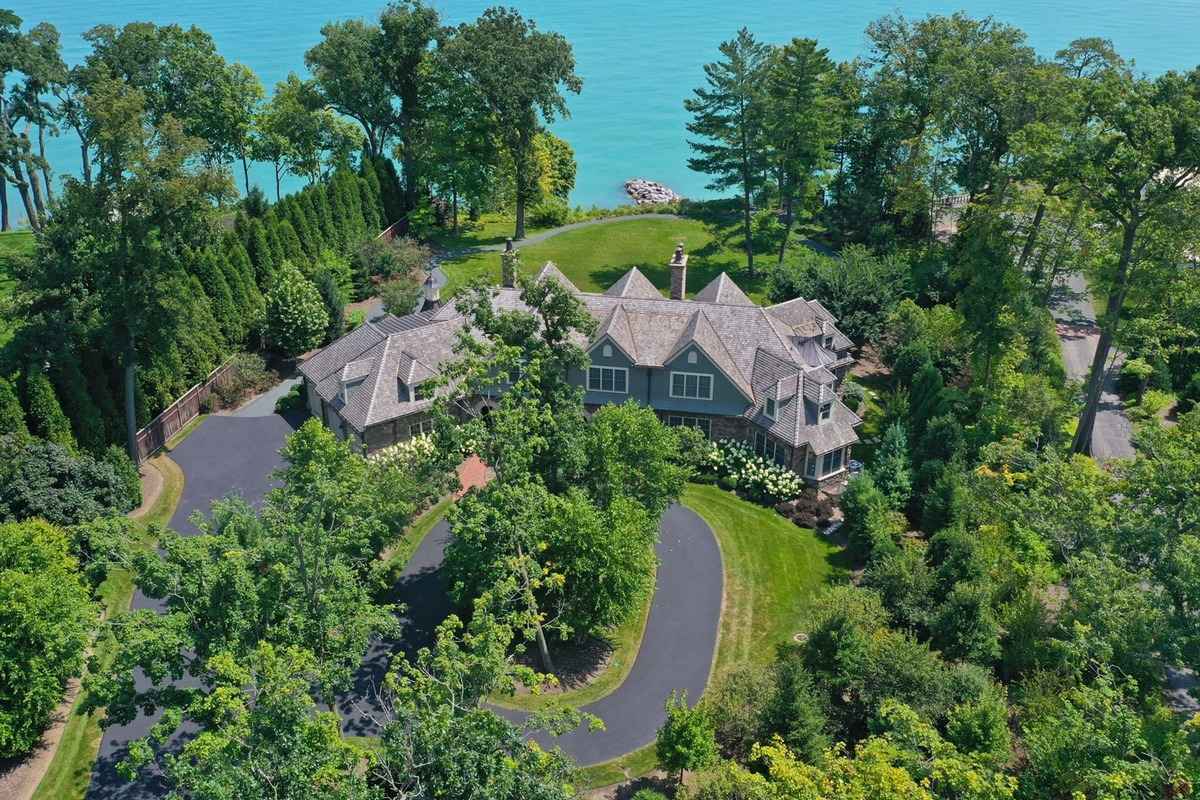 An elegant lakeside mansion surrounded by lush greenery with a circular driveway and stunning waterfront views.