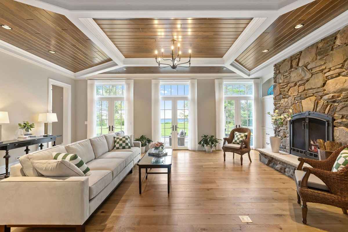 A bright and elegant living room featuring a coffered wood ceiling, a stone fireplace, and large windows overlooking lush greenery.