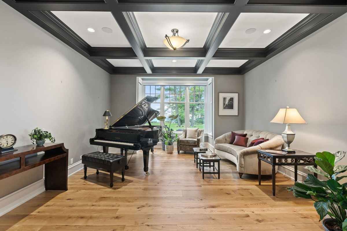 A sophisticated music room with a grand piano, coffered ceiling, and a cozy seating area near a large window.