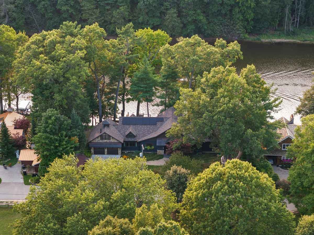 An aerial view shows a large house nestled among lush green trees beside a body of water.