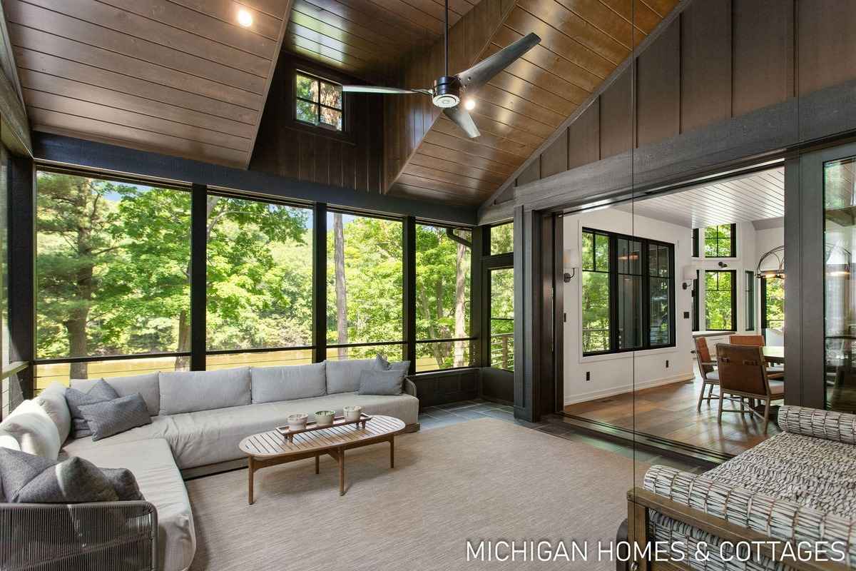 A sunroom with a vaulted ceiling, large windows, and a comfortable seating area offers a view of a wooded area.