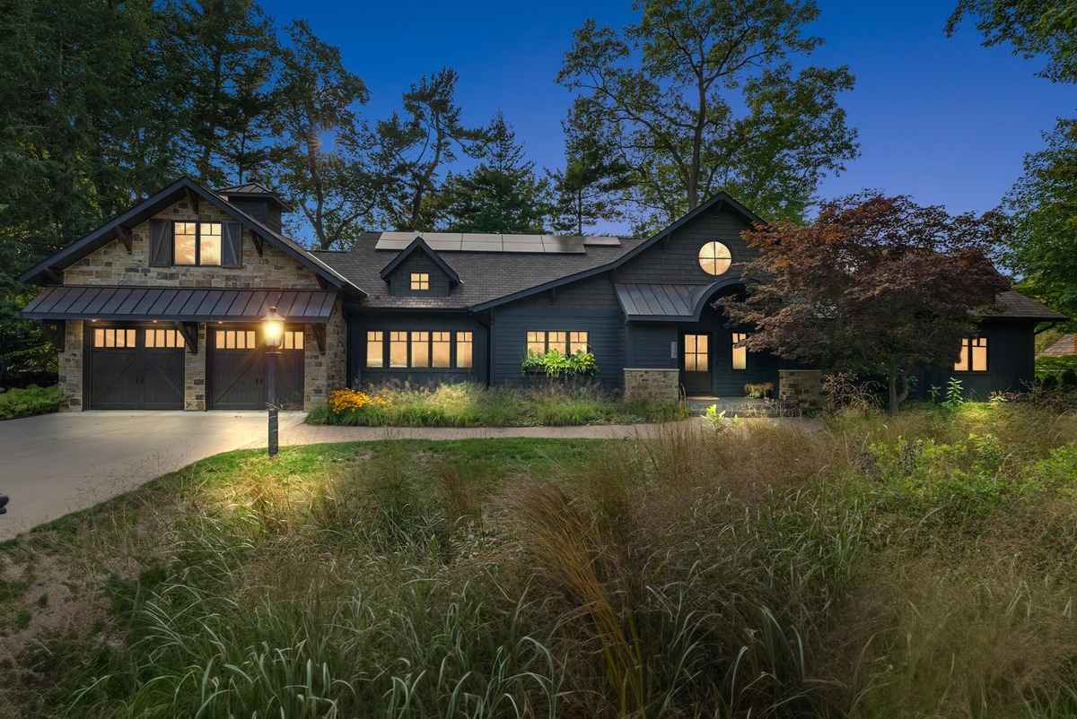 A modern, dark-colored house with a three-car garage is shown at dusk, set against a backdrop of tall grasses and trees.