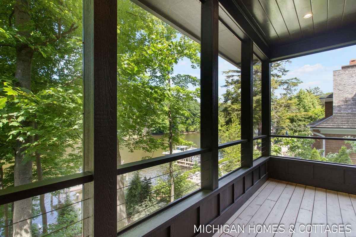 A screened porch with dark wood framing offers a view of lush green trees and a body of water.