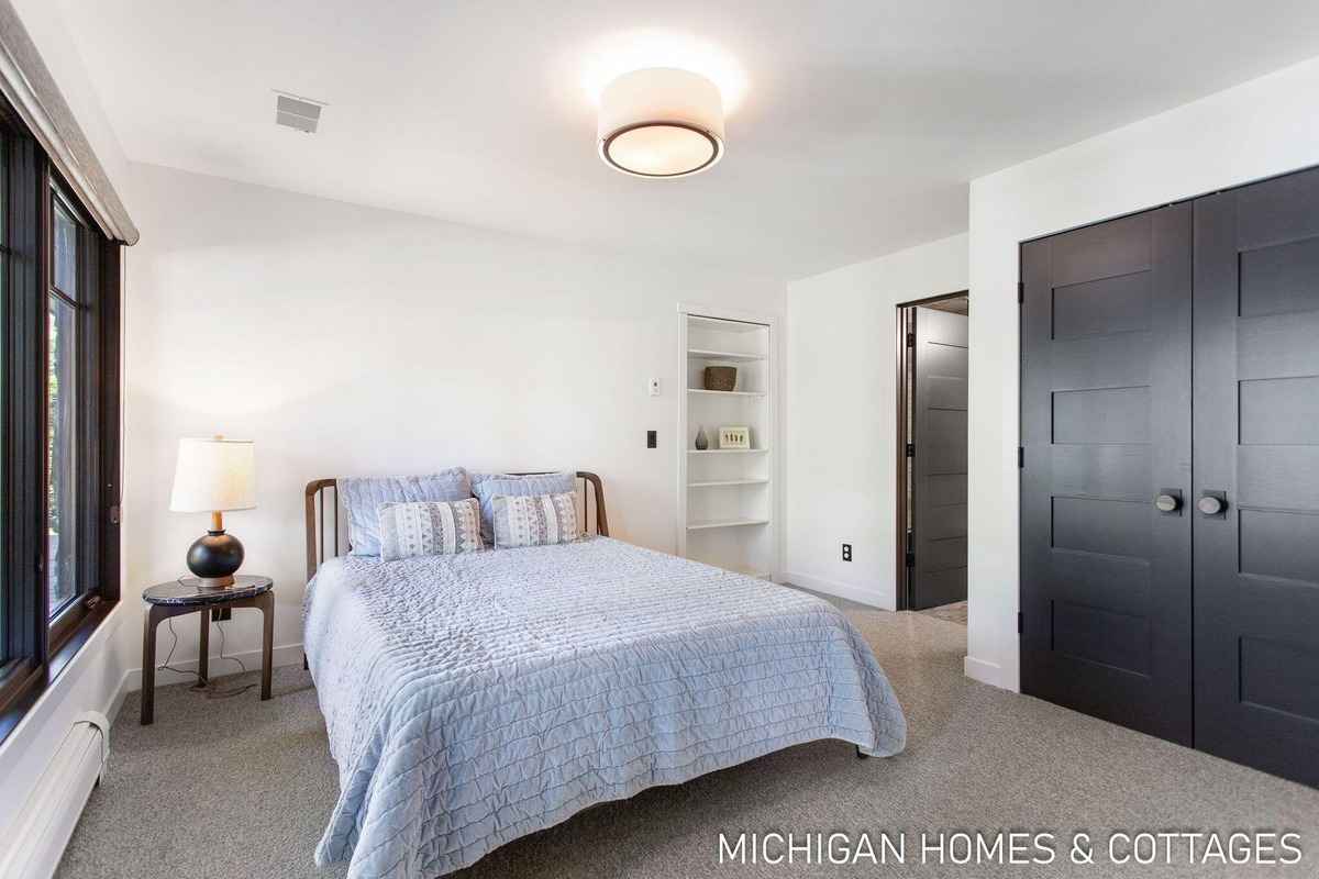 A bedroom with a light-colored wall, a bed with a light blue comforter, and dark-colored double doors is shown.