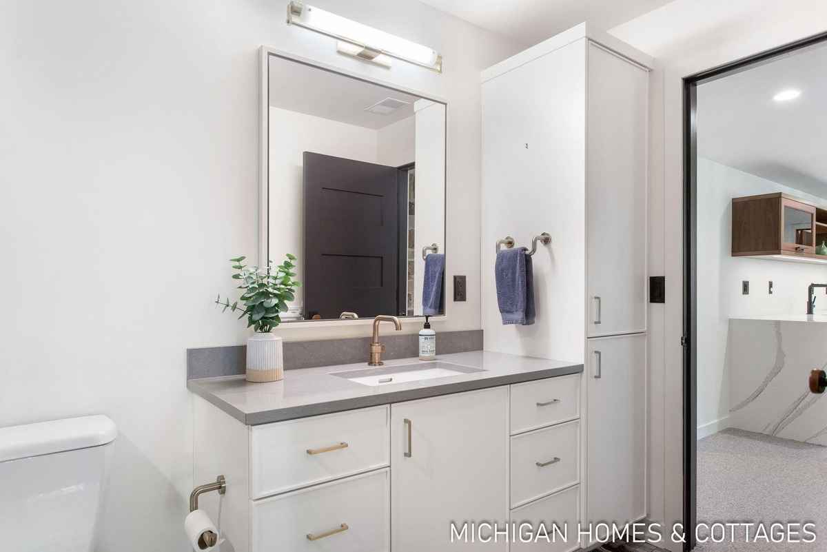 A bathroom features a white vanity with a gray countertop, a large mirror, and a glimpse of another room beyond a doorway.