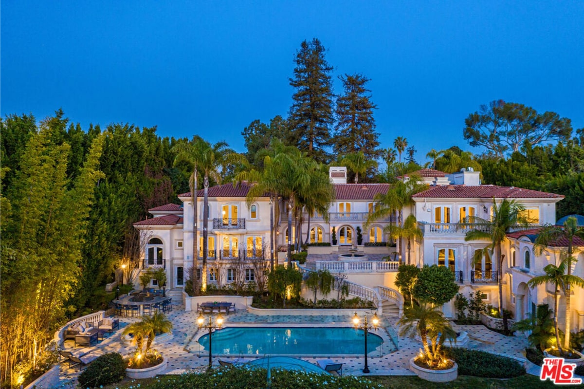 Mediterranean-style mansion at dusk. It's a multi-story building with a swimming pool in the foreground, surrounded by landscaping that includes palm trees and other lush greenery.