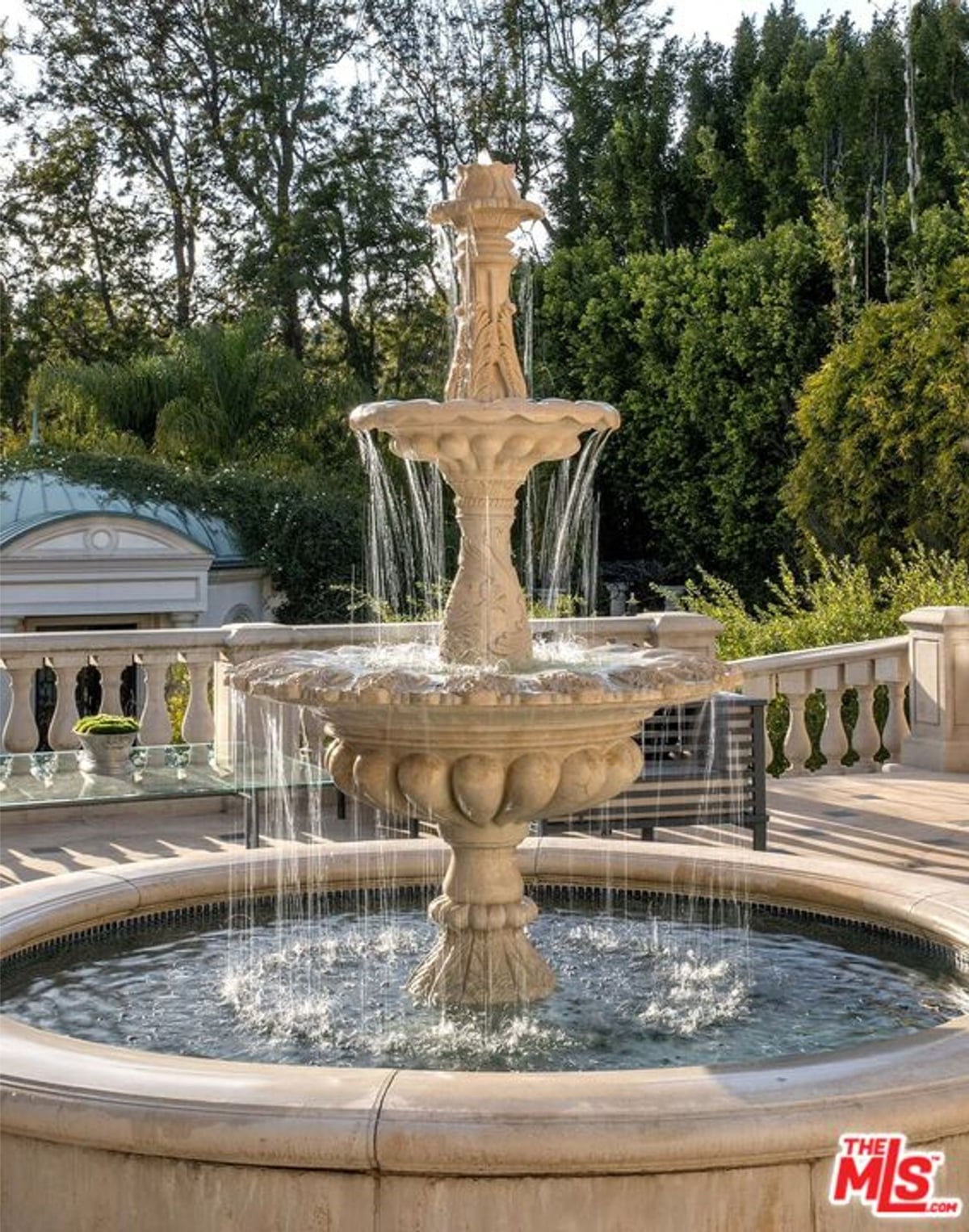 An ornate fountain in a garden setting. The fountain is light-colored stone, with multiple tiers and water cascading down. Lush greenery and trees form the backdrop.