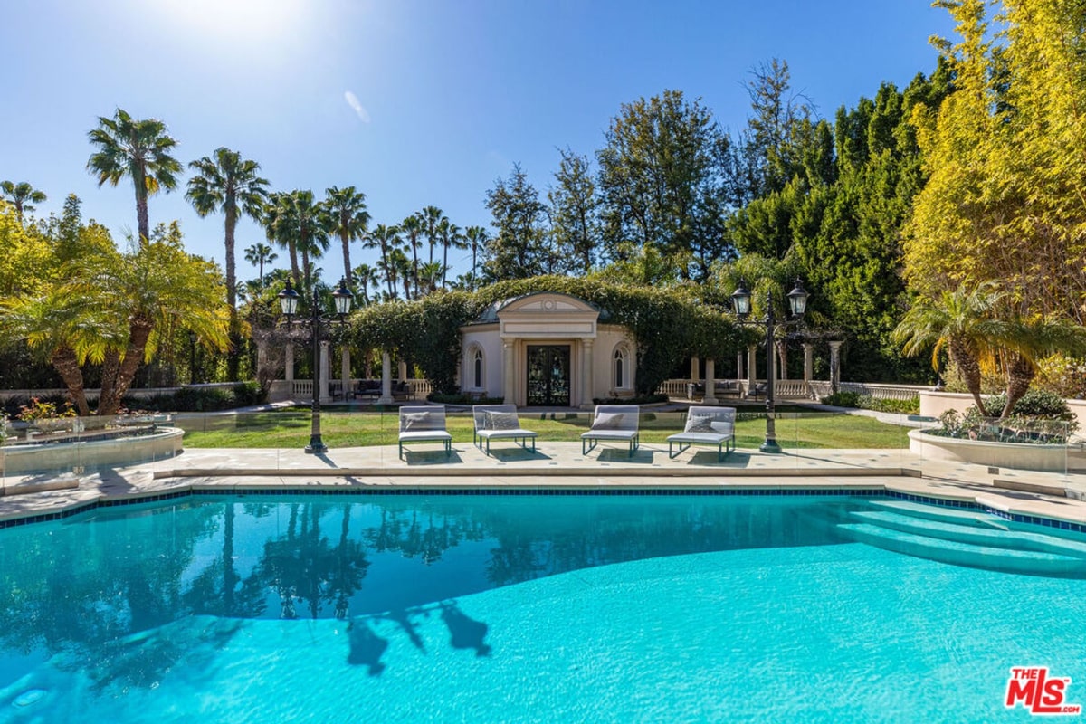 Sparkling blue swimming pool at a luxurious property. Lounge chairs are positioned on the pool deck, and in the background, there's a classical-style structure and lush landscaping with palm trees. The scene evokes a sense of opulence and resort-like living.