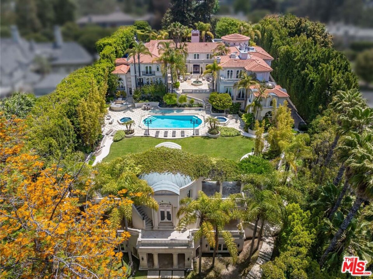 An aerial view of a large, luxurious estate. The main house is a multi-story mansion in a classic style, with a swimming pool and extensive landscaping surrounding it. The property appears to be very large and opulent.