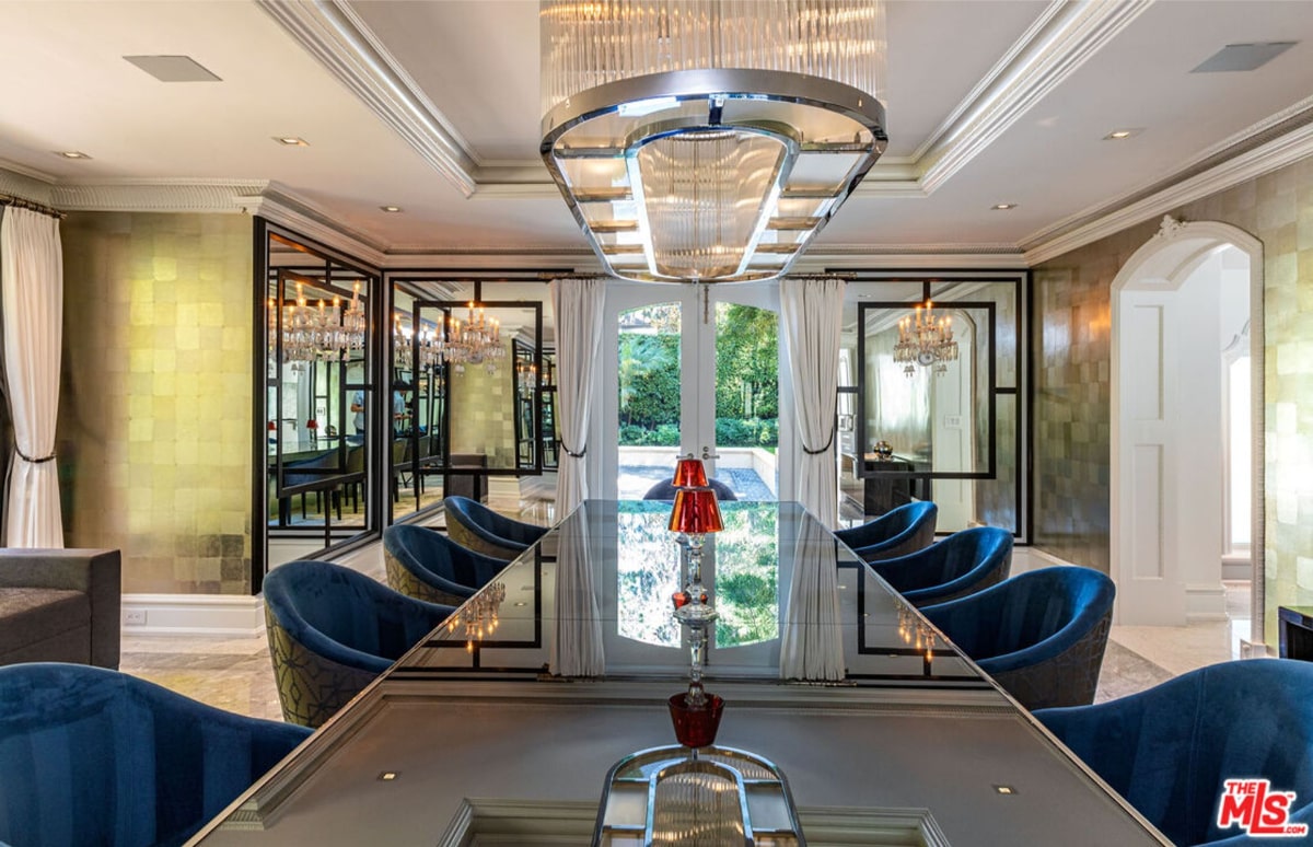 Formal dining room with a long, glass-topped table and blue velvet chairs. A large, modern chandelier hangs above the table. Large windows or glass doors offer views of the outdoors.