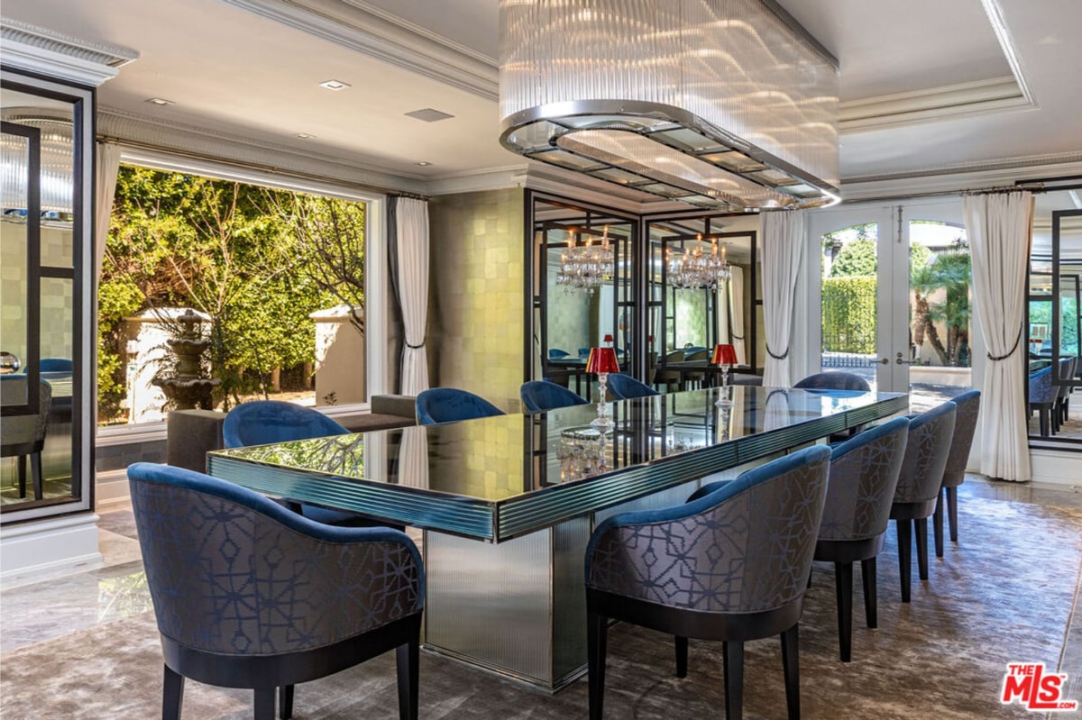 Dining room with mirrored table and elegant chairs. A dramatic chandelier is a focal point, and large windows provide a view of a garden or courtyard