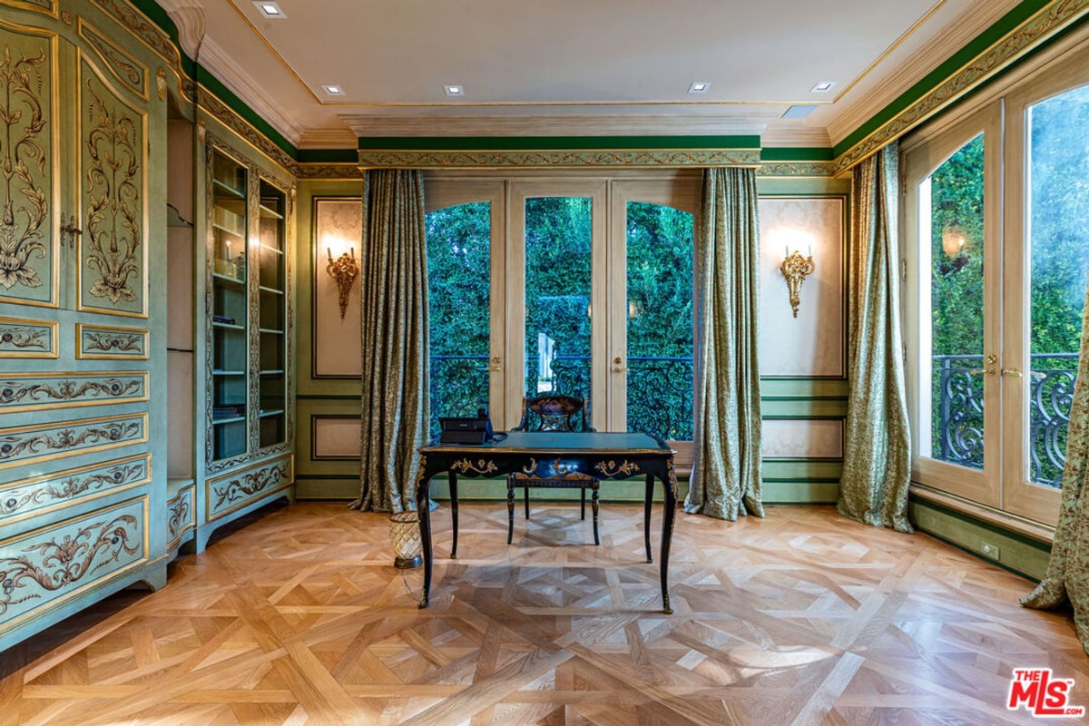 Library room with ornate, light-green cabinetry and a dark wood desk. The room features large windows with drapes, and the floor is a patterned hardwood.