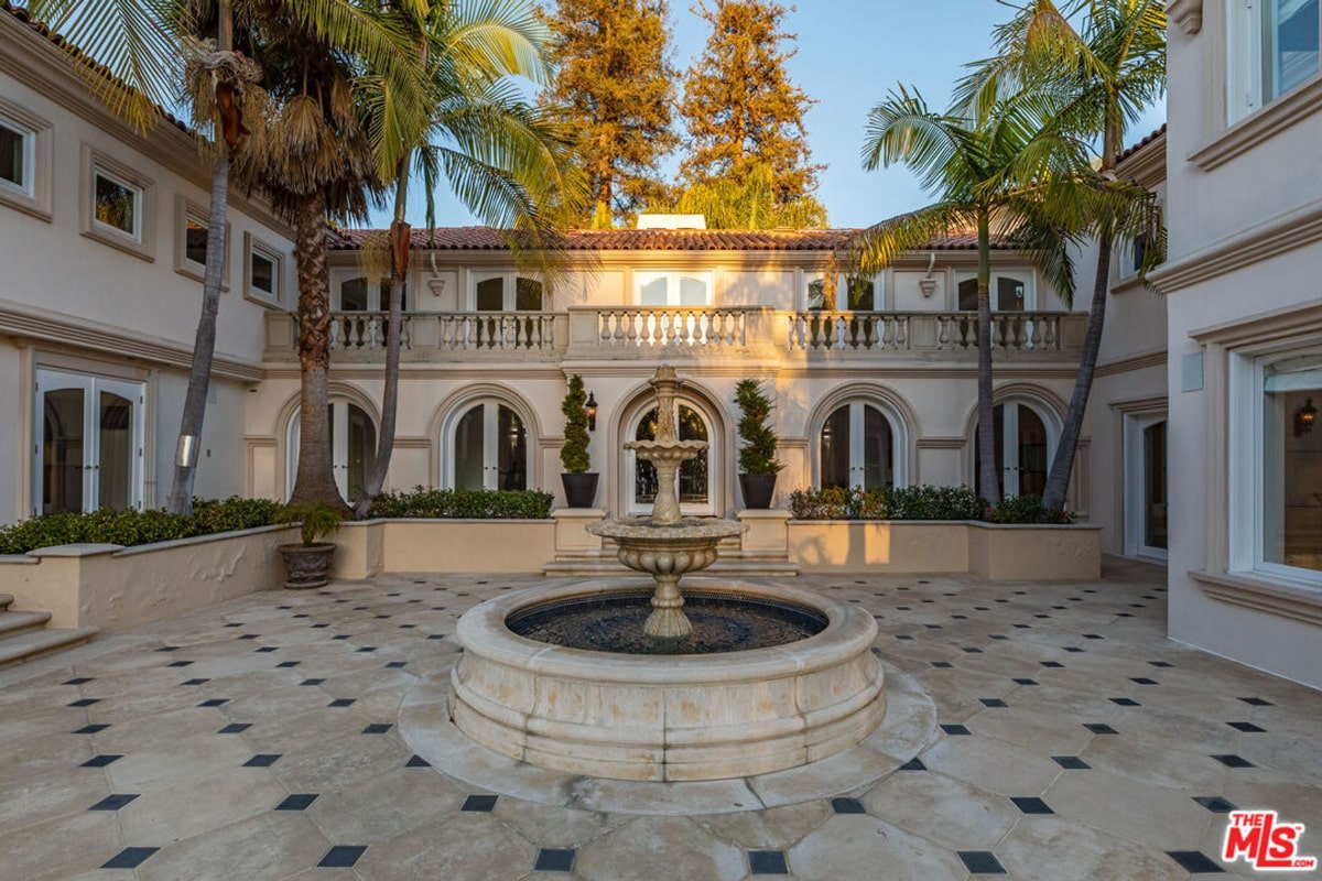 Central courtyard of a large mansion. The courtyard features a stone fountain as its centerpiece, surrounded by a paved area with a geometric tile pattern. The mansion itself is in a classical style with arched doorways and balconies, and palm trees add to the Mediterranean feel.