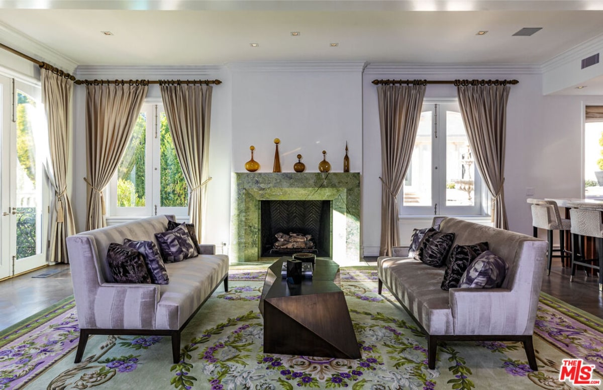 Living room with two light-colored sofas arranged around a coffee table in front of a fireplace. The room has large windows with floor-length drapes.