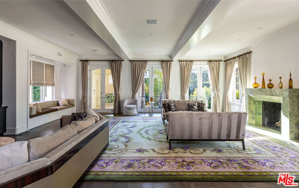 Bright living room with high ceilings and large windows. Light-colored sofas and a large area rug are arranged around a fireplace. The overall style is elegant and airy.