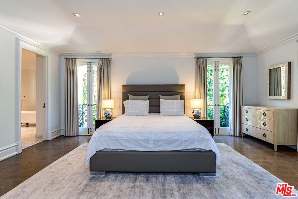 Bedroom with a large bed, a neutral color scheme, and floor-to-ceiling windows with drapes. The room has a calm, sophisticated feel. A dresser and nightstands are visible.