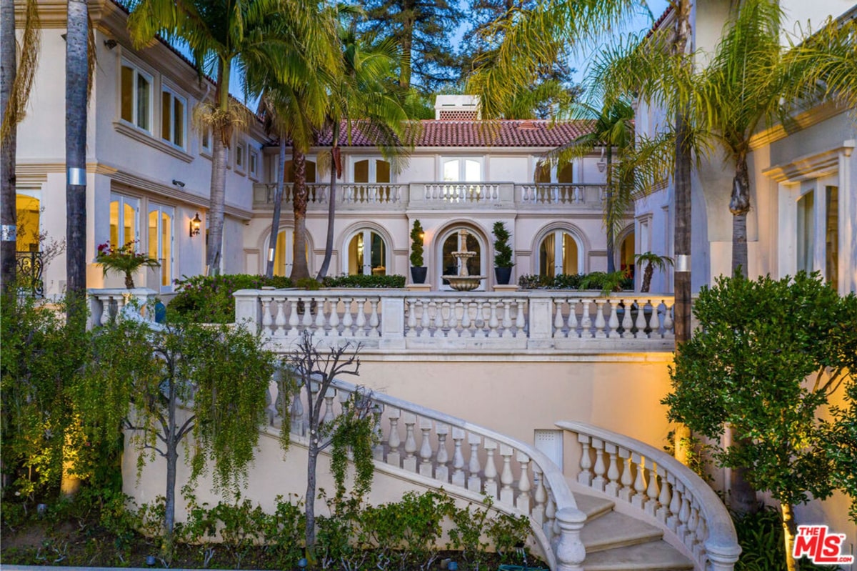 Elegant mansion at dusk or night. The architecture is classical or Mediterranean-style, with white walls and a grand staircase. Mature palm trees and landscaping surround the property.