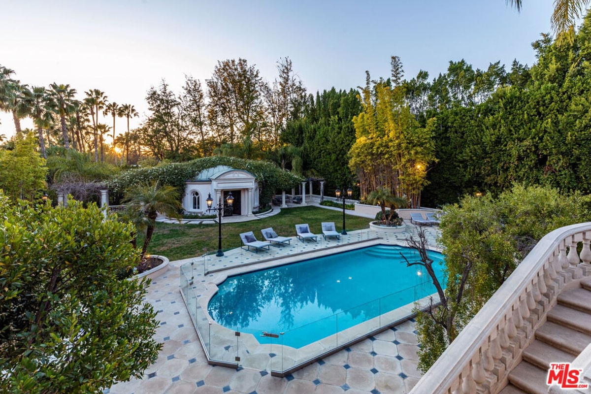 Swimming pool and patio area of a large estate. The pool is rectangular with a glass fence, and there are lounge chairs nearby. A small, classical-style structure is visible in the background, surrounded by landscaping.