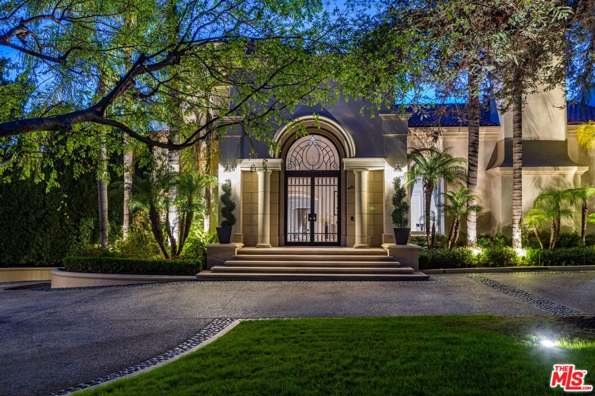 A grand home entrance at night, featuring ornate iron gates and lush landscaping.