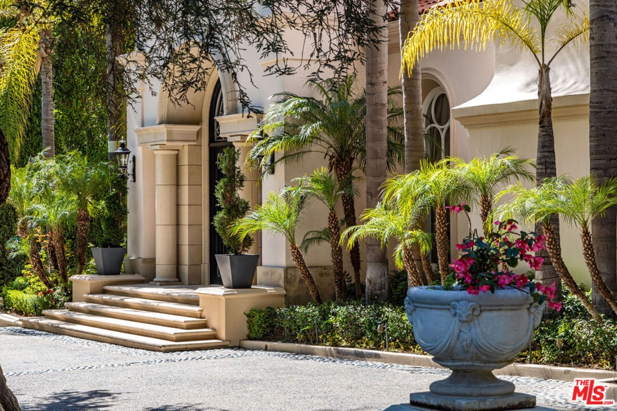 Entrance is marked by large columns and steps, and there's a large urn with flowering plants in the foreground. Palm trees and other lush landscaping are visible.