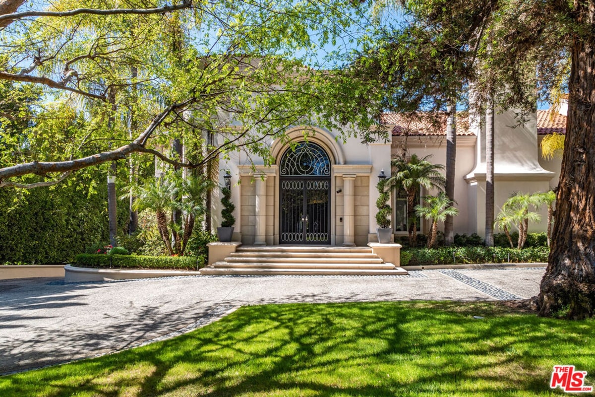 A prominent arched doorway with elaborate iron gates. The home is light-colored, and the landscaping includes mature trees and manicured lawns.