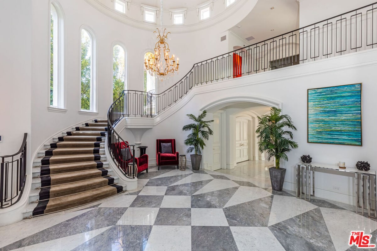  A grand foyer with a curved staircase featuring a dark runner and a black metal railing. The floor is a geometric pattern of gray and white tiles. There's a large chandelier and some potted plants, along with seating in a red and neutral color palette.