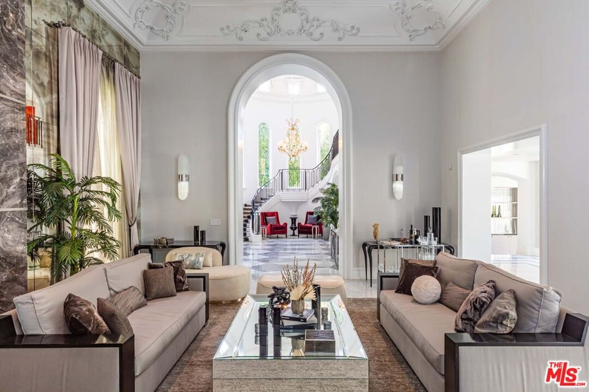  Spacious living room with a neutral color palette. Two large sofas face each other across a large coffee table. An arched doorway in the background leads to another area with a grand staircase and red chairs, suggesting a luxurious and elegant home.