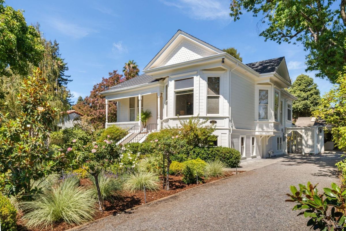 White exterior house with a sloped roof surrounded by lush landscaping and a gravel driveway.