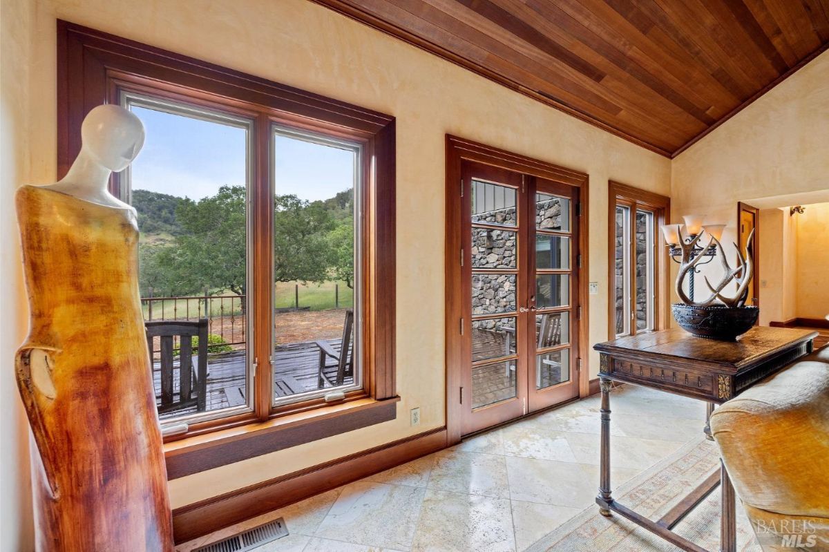 A cozy interior space featuring large windows with a view of the outdoors, French doors, a wooden ceiling, and elegant decor elements.