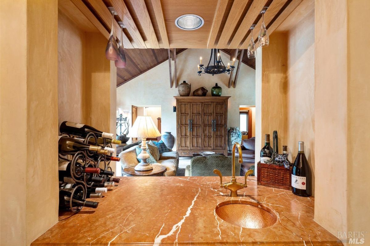 Stylish wet bar with marble countertops, wine racks, and a copper sink, leading to a view of the living room.