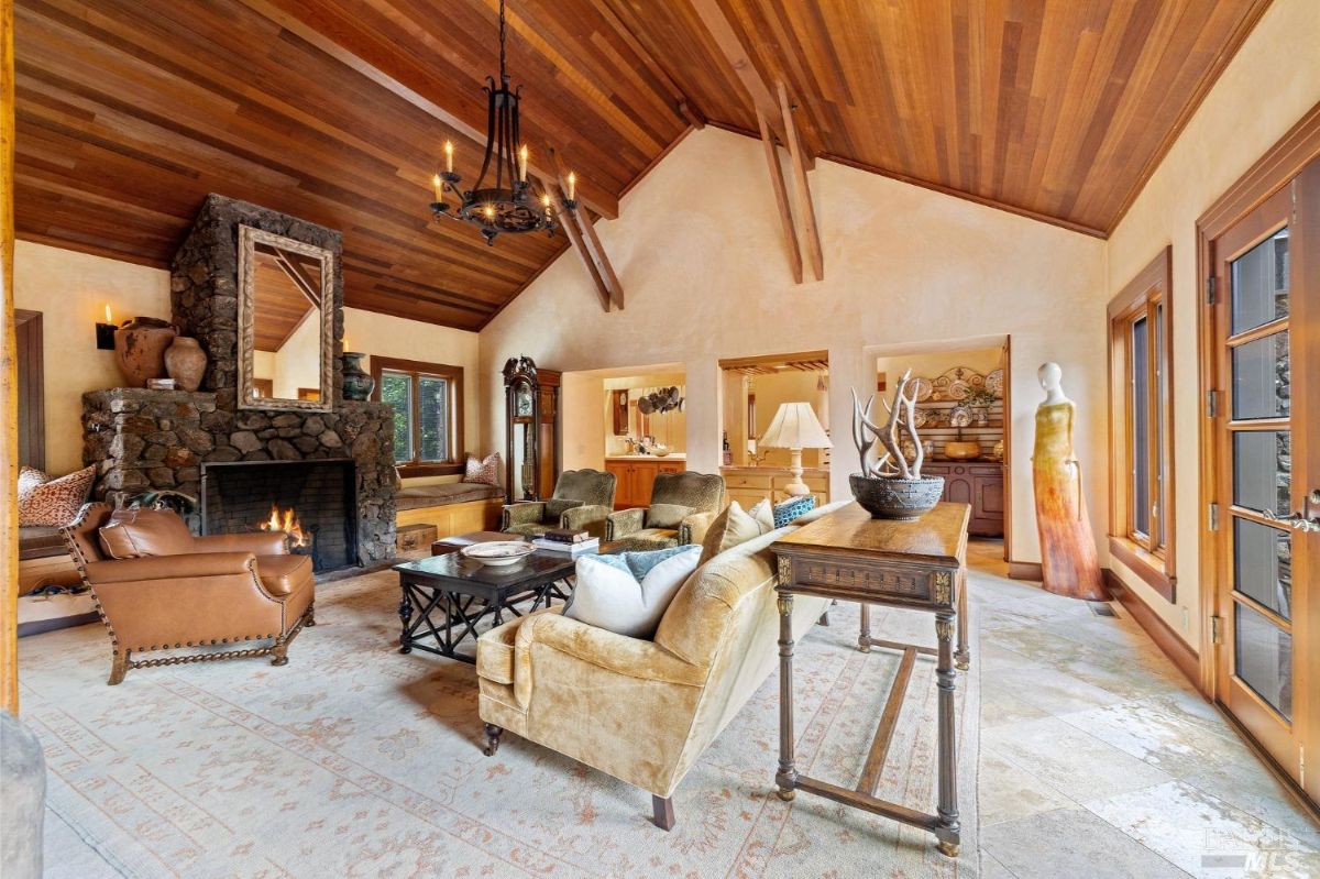 A grand living room featuring a stone fireplace, vaulted wooden ceiling, and a mix of vintage and modern furniture.