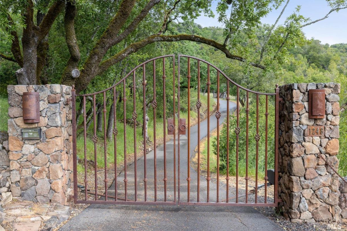 A gated entrance made of wrought iron and stone pillars, leading to a long driveway flanked by trees and natural surroundings.