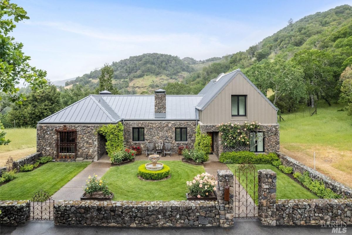 A rustic stone home with a symmetrical courtyard, lush landscaping, and a central fountain surrounded by vibrant greenery and flowers.