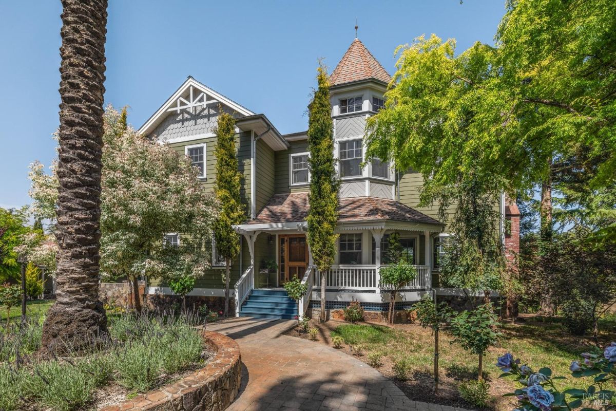 Victorian-style house with a green exterior, white trim, and a turret feature surrounded by lush landscaping.