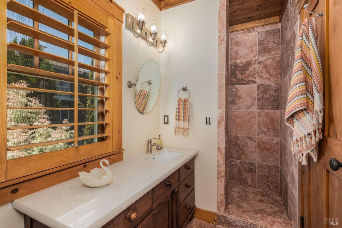 Bathroom has a single vanity, a walk-in shower with brown tile, and a window with wooden shutters.